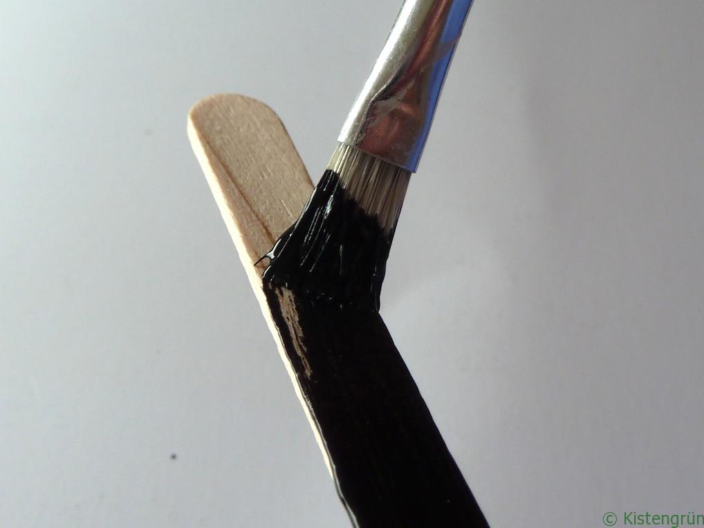 Mit einem breiten Pinsel wird ein Holzstab mit schwarzer Tafelfarbe bemalt.